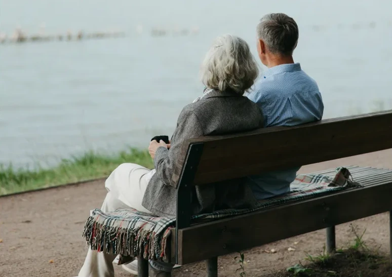 The back view of an elderly couple sitting close together on a wooden park bench. They are looking out peacefully over a calm body of water.