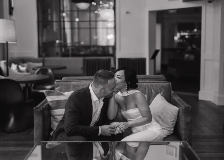A romantic black and white photograph of a well-dressed couple sitting closely on a leather sofa in an elegant lounge.
