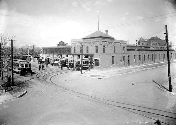 Historic black-and-white image of a street intersection featuring a trolley, buildings, and utility poles in an early 20th-century city setting.