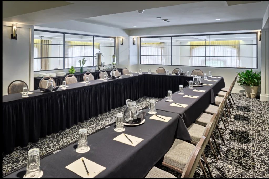 A conference room is set up with a U-shaped table covered in black cloths, with chairs, notepads, and water glasses ready for a meeting.