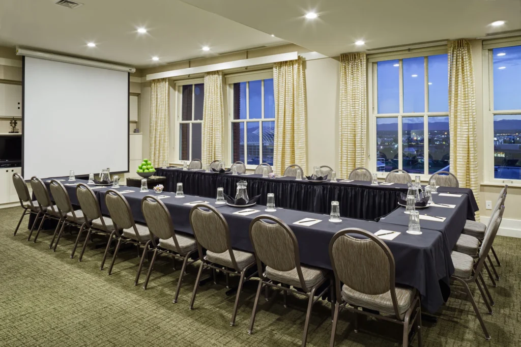 A conference room with a U-shaped table facing a projector screen, with large windows showing a city view at dusk.