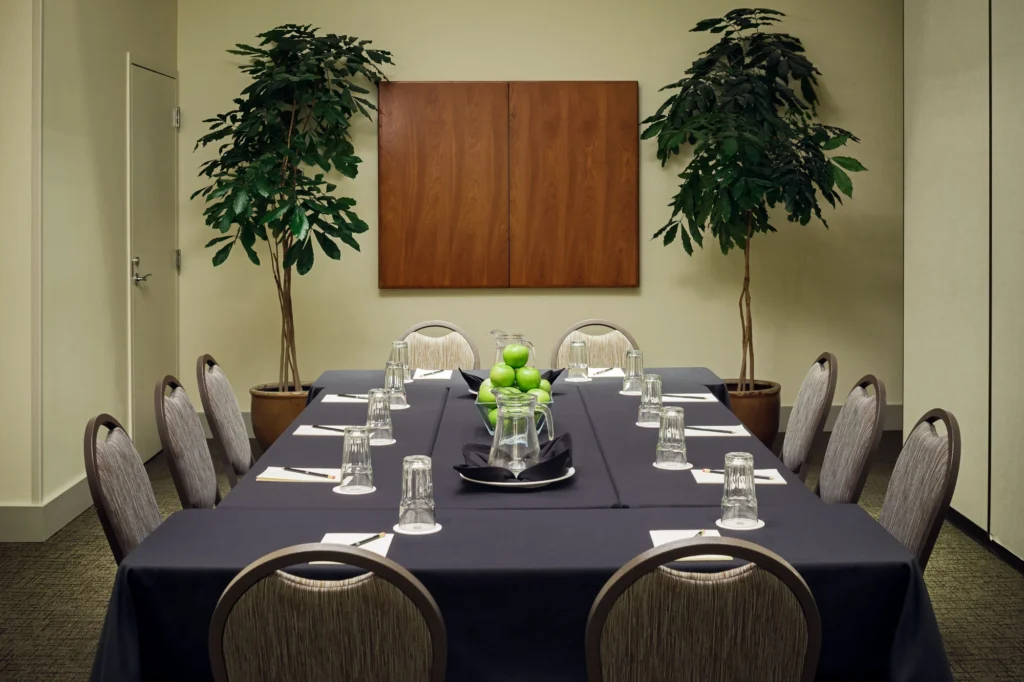 A rectangular conference table with a dark tablecloth is set with glasses and notepads for ten people, with a bowl of green apples in the center.