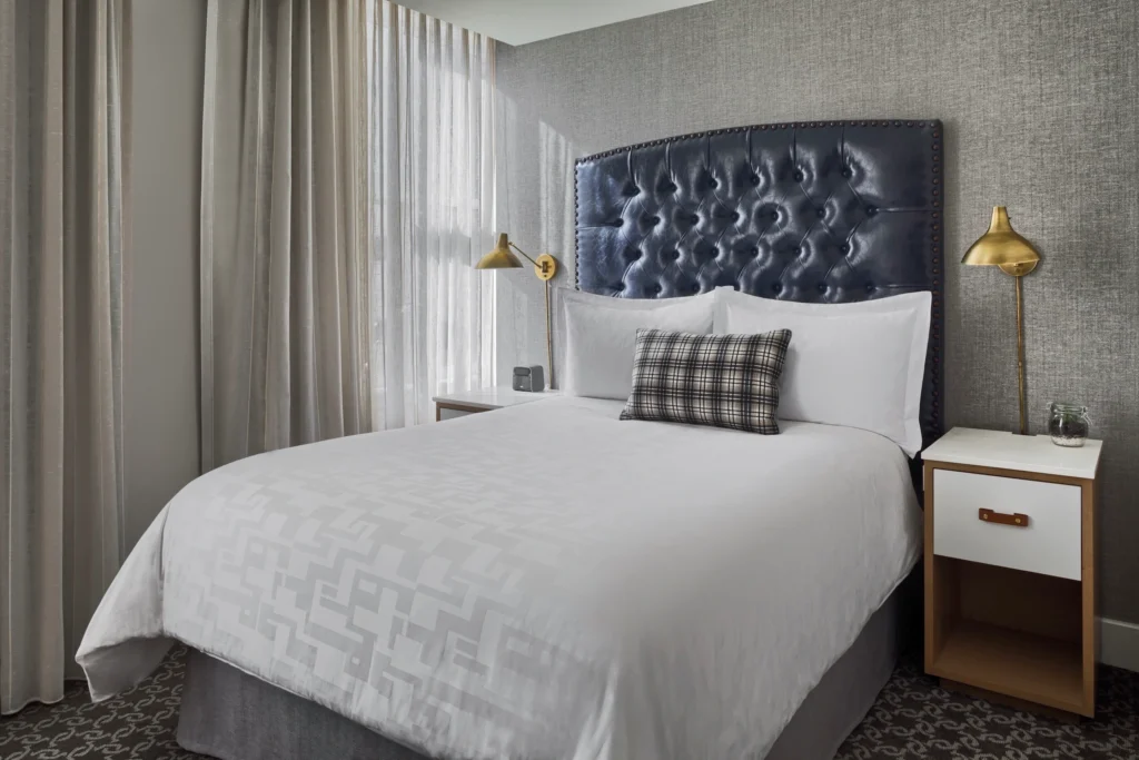 A stylish hotel room featuring a neatly made bed with a patterned duvet and plaid accent cushion, soft curtains, and a modern bedside lamp.