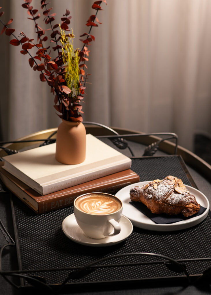 a latte and croissant on a tray next to a small stack of books and flowers in a vase