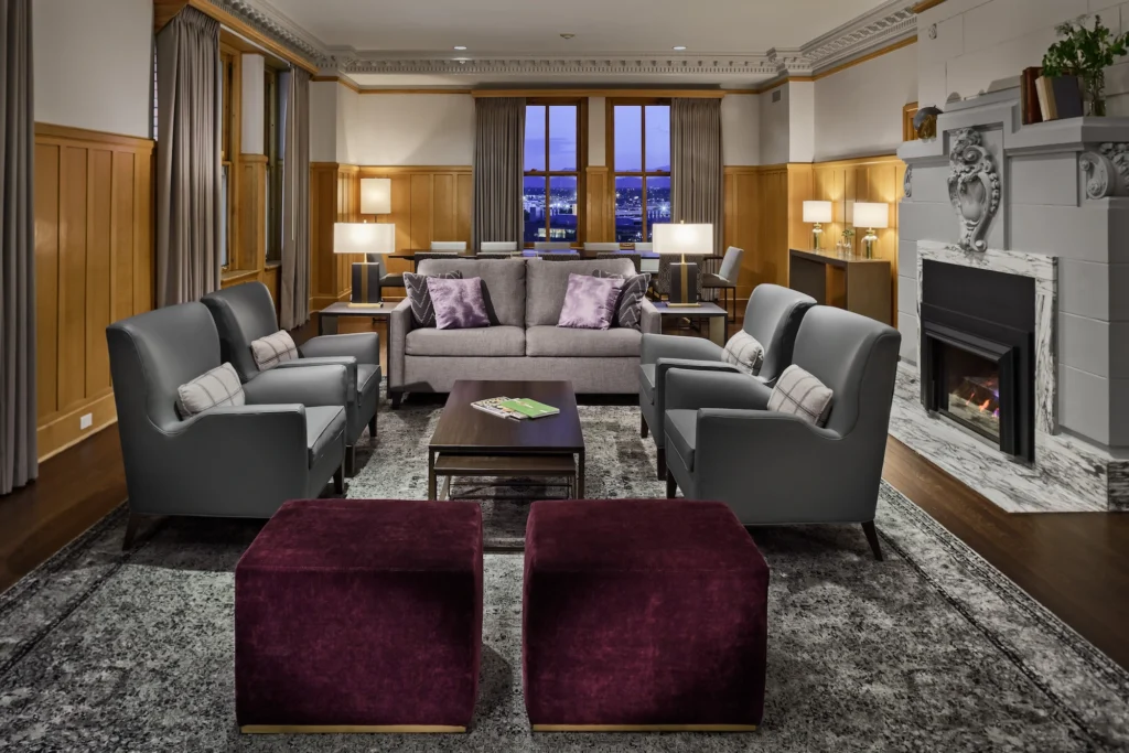 A stylish lounge with grey armchairs and a sofa, plush purple ottomans, warm wood accents, and a cozy fireplace, illuminated by soft lamps.