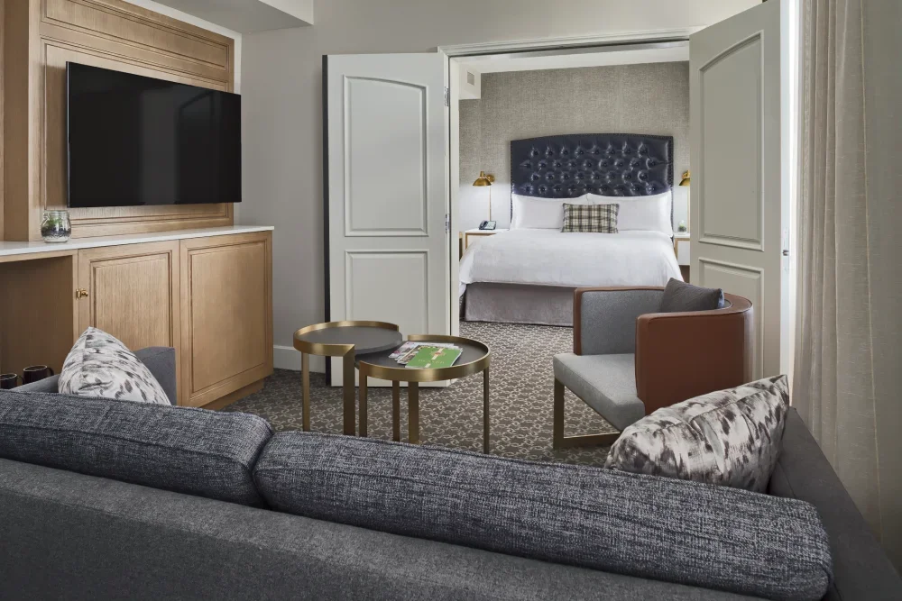 A hotel junior suite with a view from the living area into the separate bedroom. The living room in the foreground features a gray sofa and a modern armchair.