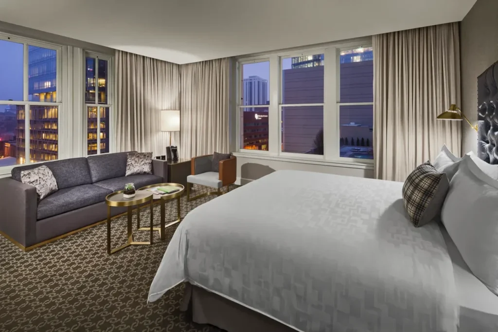A modern hotel room featuring a king bed, comfortable seating area, large windows, and elegant decor overlooking the city.