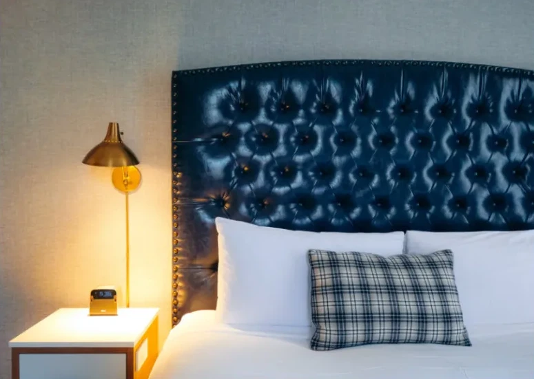 A close-up of a well-made hotel bed featuring a large, glossy navy blue tufted headboard. The bed is adorned with white pillows and a blue and white plaid decorative pillow.