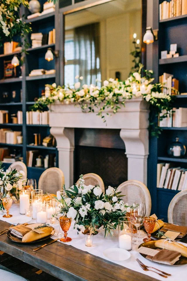 A close-up of an elegant dining table set with white floral arrangements and candles, in front of a grand fireplace framed by blue bookshelves.